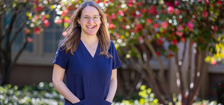 A woman with brown hair and glasses wearing navy blue medical scrubs stands outside near flowering trees. 