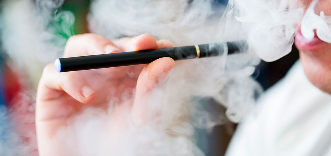 A hand holds a black e-cigarette surrounded by a cloud of white smoke