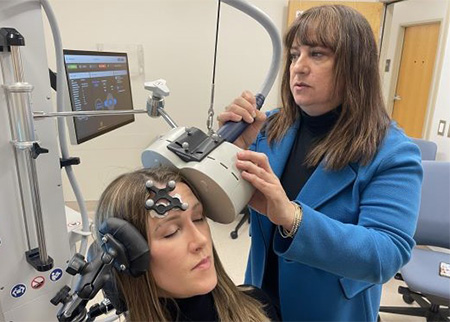 Una mujer con un dispositivo de seguimiento negro en la frente está sentada mientras otra mueve una bobina magnética beige cerca de su cabeza.