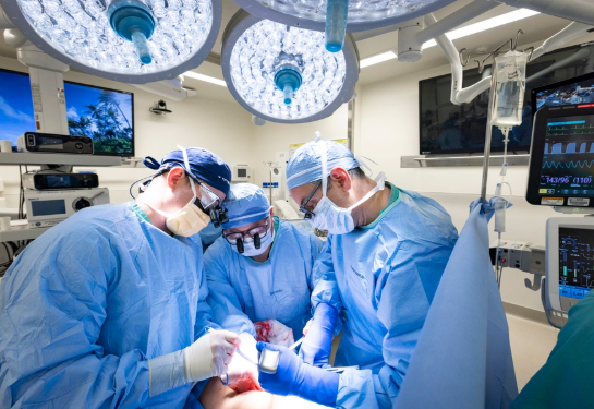 Three surgeons wearing blue surgical scrubs stand around a patient lying on a surgery bed.