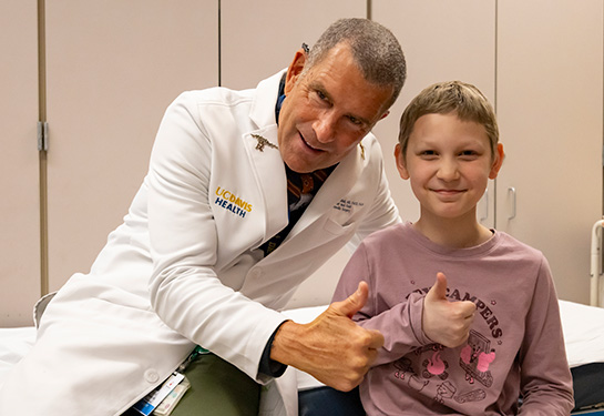 Man in white coat sits next to girl in pink shirt. Both give thumbs up.