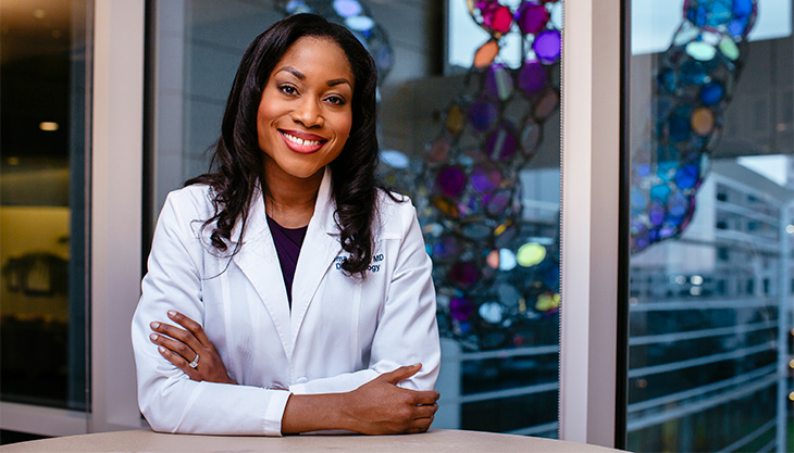 Dr. Oma Agbai is in a white coat. She stands smiling with crossed arms in front of her and a glass art piece behind.