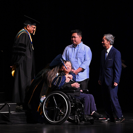 A young woman in a blue gown kisses the cheek of a woman in a wheelchair, surrounded by three men who are standing