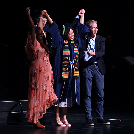 A young woman in a blue gown stands between a man and a woman on stage as they hold hands and raise their arms into the air 