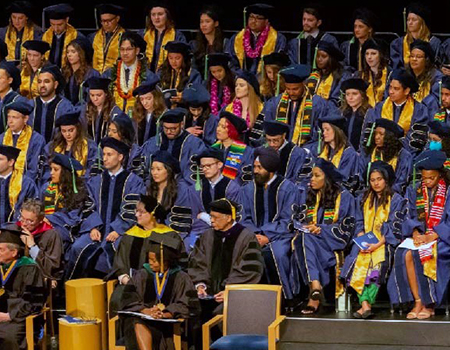 Dozens of students sit on stage wearing black caps, yellow stoles and blue gowns
