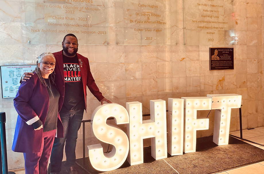 Jann Murray-García and Aron King stand beside a lighted sign that reads SHIFT