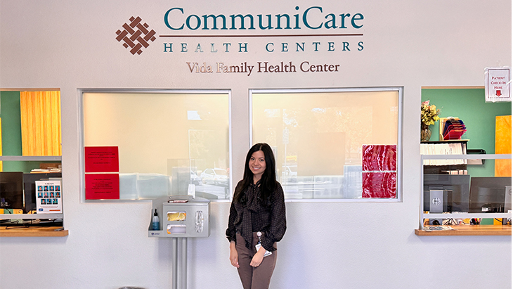 A woman with brown hair stands in the lobby of a clinic with a sign behind her that reads: “CommuniCare Health Centers, Vida Family Health Center.”