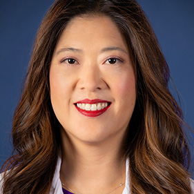 A woman with long brown hair, wearing a white doctor’s jacket and purple shirt underneath smiles for a portrait.