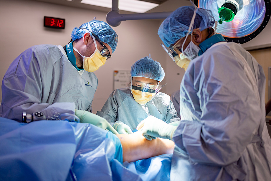 Three people in medical scrubs perform a procedure on a patient’s knee in an operating room. 