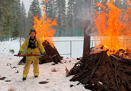 A firefighter stands near several piles of burning brush.