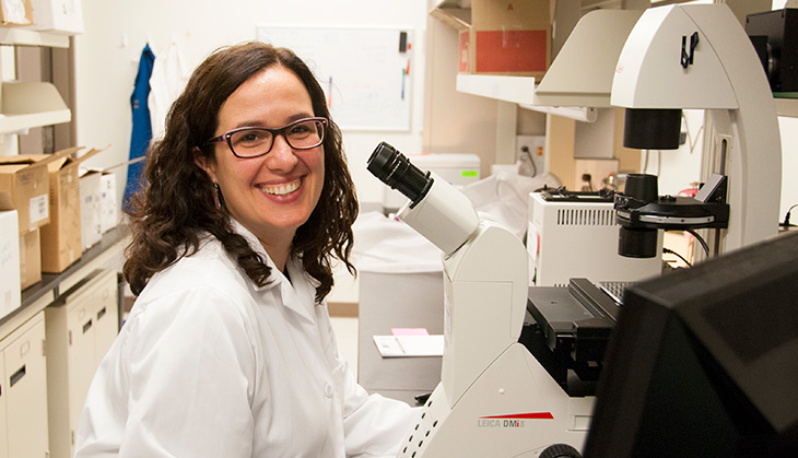 Professor Anna La Torre is smiling and looking at the camera. She is wearing eyeglasses and white lab gown is sitting in front of a microscope.