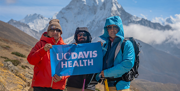 Three men with cold weather jackets and hats holding UC Davis Health flag with snowy mountain in the background