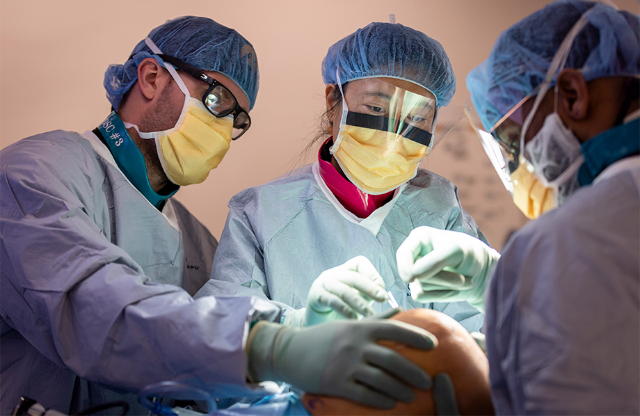 Three surgeons in medical scrubs gather around a patient's knee as they perform a surgery in an operating room. 