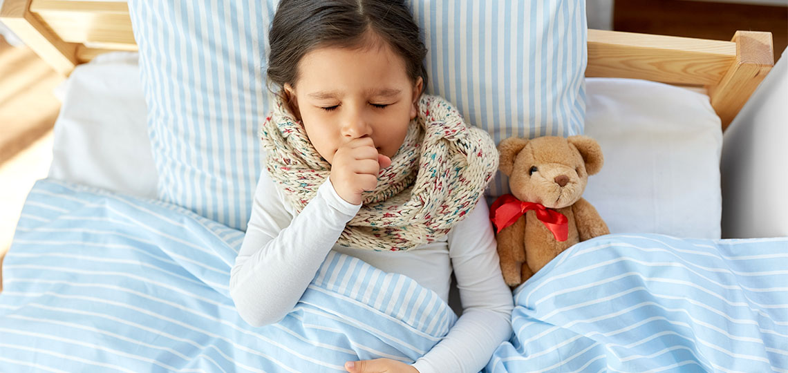 Girl lies in bed coughing, with her hand covering her mouth. A teddy bear is next to her.