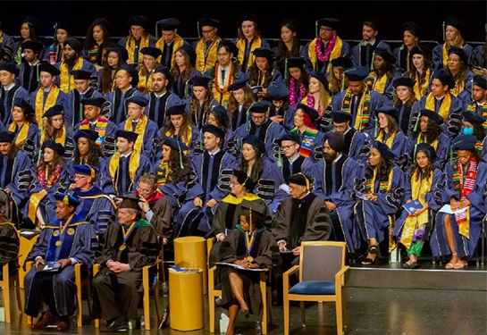 Dozens of medical students sit on stage wearing black caps, yellow stoles and blue gowns