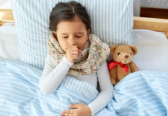Girl lies in bed coughing, with her hand covering her mouth. A teddy bear is next to her.