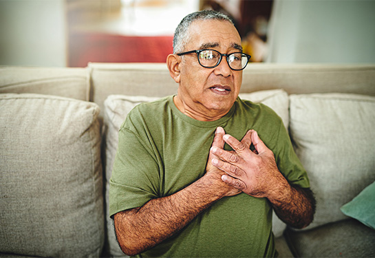 Un hombre de edad avanzada, de cabello gris y sentado en un sillón, detiene su pecho con sus dos manos
