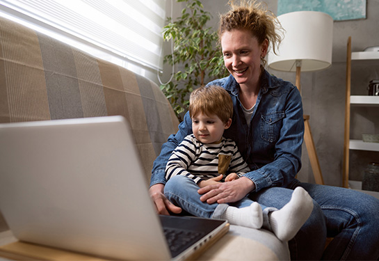 A mother and son sit on a couch, looking at a laptop, which is open beside them, as they engage with someone on a video call.  