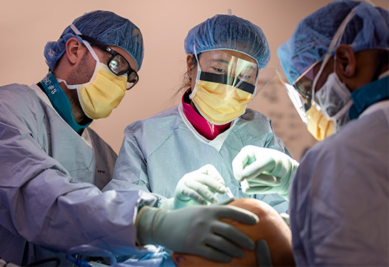 Three surgeons in medical scrubs gather around a patient&#x2019;s knee as they perform a surgery in an operating room. 
