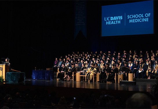 Stage with hundreds of people sitting in row wearing caps and gowns with screen in back reading UC Davis School of Medicine.