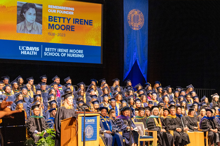 Graduates sit on risers on stage while woman speaks from podium; screen with photo honoring co-founder Betty Irene Moore shows overhead