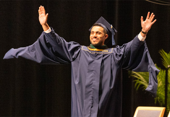  Man in graduation robe walking with arms outstretched and smiling as he walks across stage