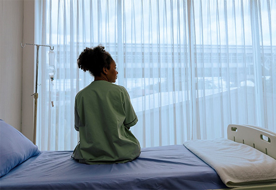 A woman sits on a hospital bed in front of a large window. 