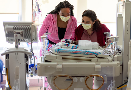 Two women stand over a baby in a bed in the NICU.