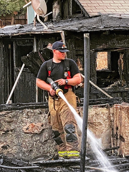 A man in firefighting gear stands amidst a burned home while spraying a thick stream of firehose water on the ground