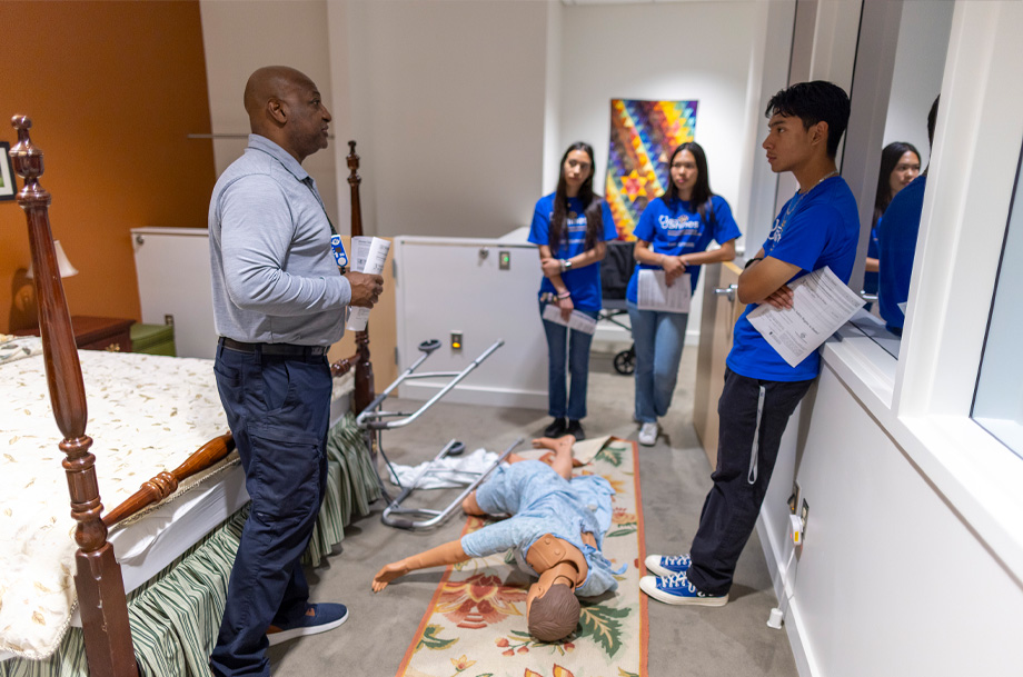 Man, left stands in apartment simulation speaking to students, right, with manikin and overturned walker on floor in front of bed