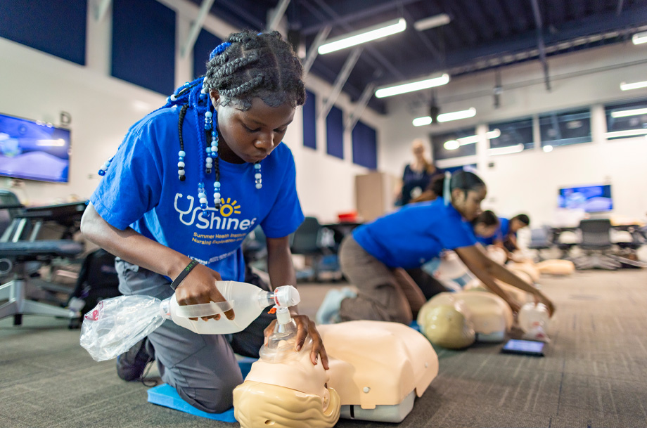Student on knees uses bag valve mask on mannequin during CPR training.