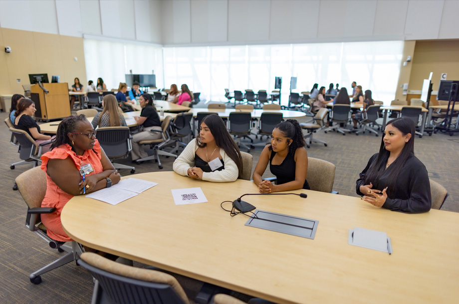 woman, left, sits at table talking with students surrounding sides in full classroom