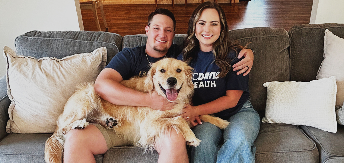 A husband and wife sit together on a couch with a large dog resting on the man’s lap