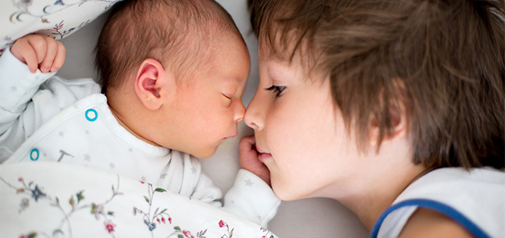 A young boy touches his nose to his infant sibling’s nose as the two cuddle in a bed.