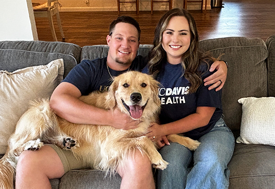 A husband and wife sit together on a couch with a large dog resting on the man’s lap