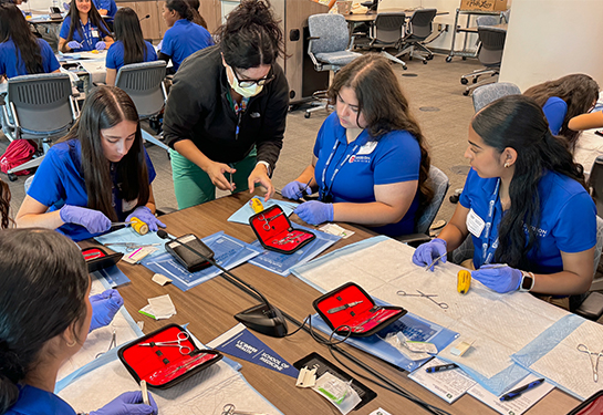 UC Davis Health surgeon Anamaria Robles leans over a table to show four teenage girls in blue polo shirts how to suture a banana