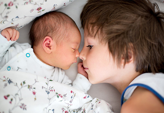 A young boy touches his nose to his infant sibling’s nose as the two cuddle in a bed.