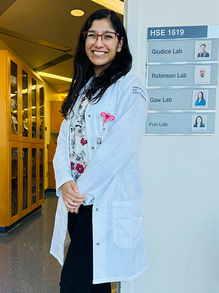 A young woman with dark hair and a white lab coat stands in a hallway next to a small directional sign that states “Giudice Lab”