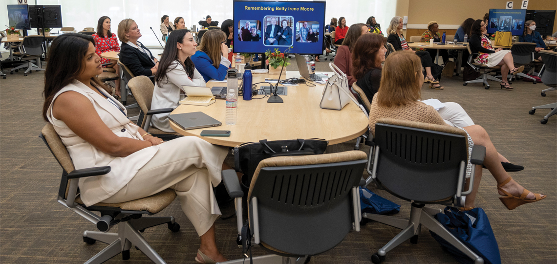 Room of fellows sitting at tables and looking off to side at speaker (not visible); screen at end of table shows photo of Betty Irene Moore and reads “Remembering Betty Irene Moore”