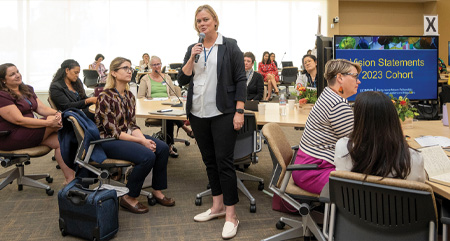 Woman stands speaking into microphone while fellows seated at tables around her look and listen