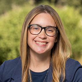 A woman with long brown hair, wearing a blue jacket and shirt and glasses smiles while standing outside.