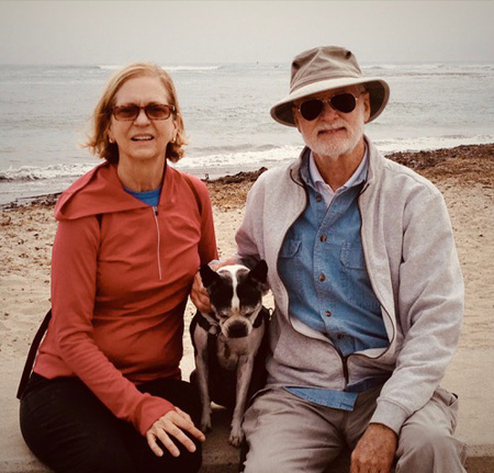 Couple sits on rock at the beach, each with an arm around their dog in the middle of them