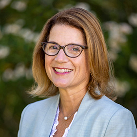 A woman with brownish-blonde hair wearing a light blue suit jacket smiles in front of greenery. 