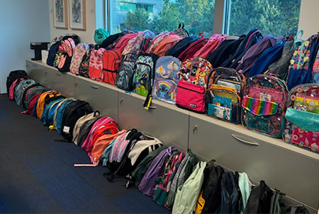 Colorful backpacks are lined up in two rows along a windowsill and the carpet in front of a large window. 