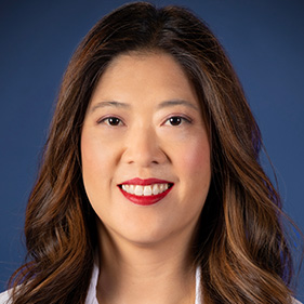 A woman with long dark hair wears a purple shirt and white doctor's coat. 