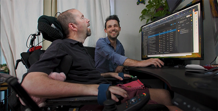 Two men looking at a computer screen: One in a wheelchair and the other looking at that man.