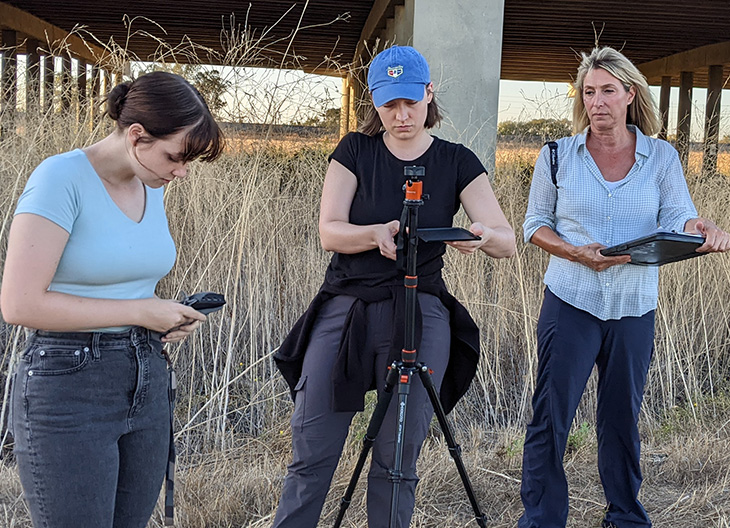 In a field, three adults set up audio and video equipment to monitor wildlife. 