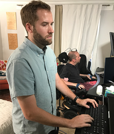 Nicholas Card standing in front of a screen. He has his left hand on a keyboard. In the background, Casey is in his wheelchair.