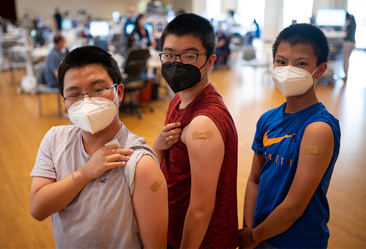 Three children stand next to each other and show on their arms the places where they were vaccinated. They are all wearing masks.