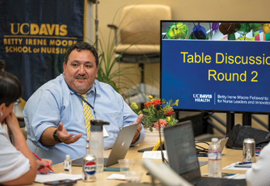 Man sits next to monitor that says &#x201c;Table Discussion Round 2&#x201d; and gestures with his right hand while talking to woman sitting next to him; Betty Irene